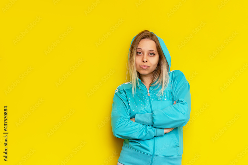 Sporty pretty confident caucasian young blonde woman in a turquoise sports hooded jacket stands with her arms crossed isolated on a bright color yellow background. Independence concept Sporty pretty confident caucasian young blonde woman in a turquoise sports hooded jacket stands with her arms crossed isolated on a bright color yellow background. Independence concept