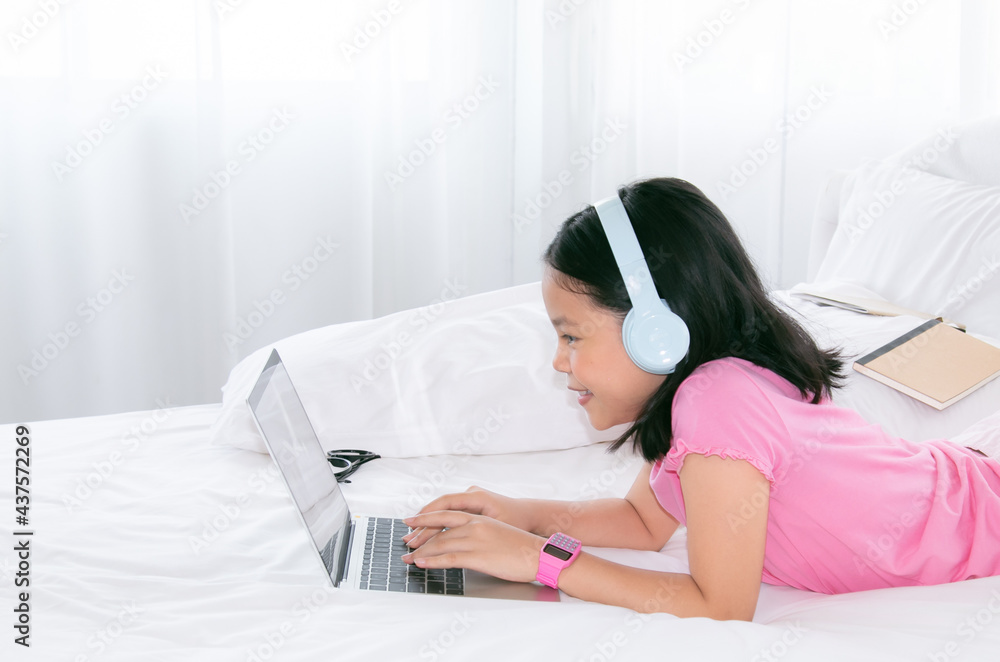 Adorable Asian girl concentrate online education class on laptop, kids lie down on bed watching game or social media connect to friends, smart school girl study e-learning using headphone technology Adorable Asian girl concentrate online education class on laptop, kids lie down on bed watching game or social media connect to friends, smart school girl study e-learning using headphone technology