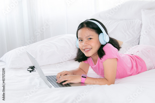 Adorable Asian girl concentrate online education class on laptop, kids lie down on bed watching game or social media connect to friends, smart school girl study e-learning using headphone technology