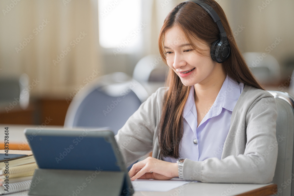 Young Asian woman college student with wireless headphones watches and listens to the lecturer on tablet and writes on the notebook to study online by herself in classroom Young Asian woman college student with wireless headphones watches and listens to the lecturer on tablet and writes on the notebook to study online by herself in classroom