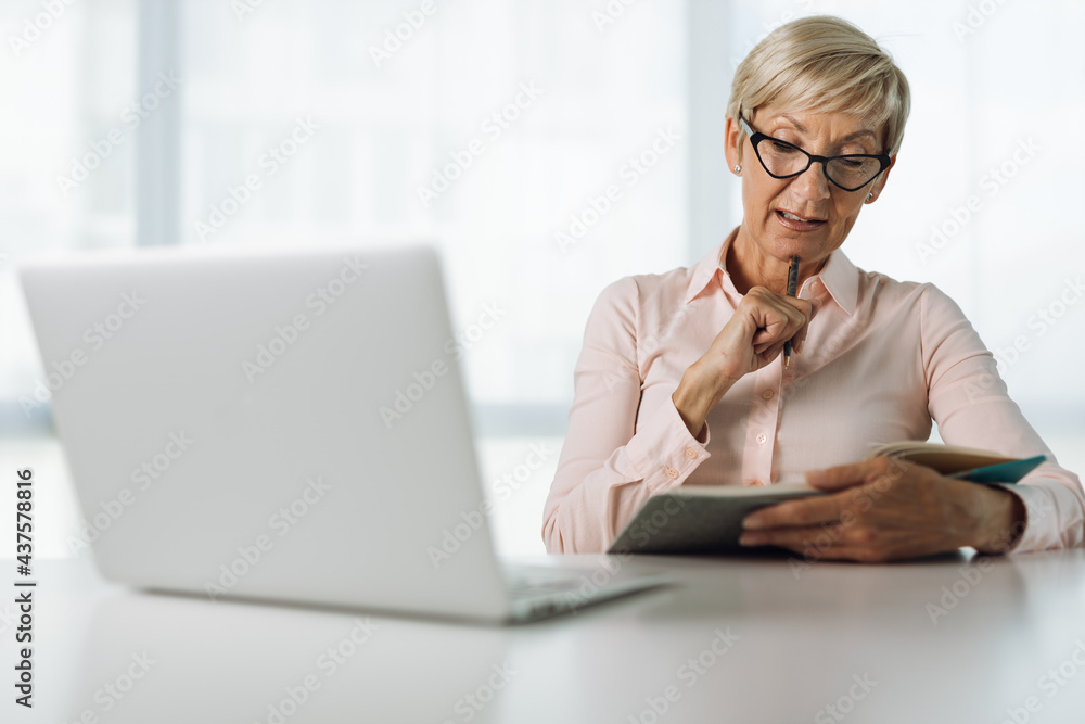 Mature businesswoman working on laptop and taking notes in the office Mature businesswoman working on laptop and taking notes in the office