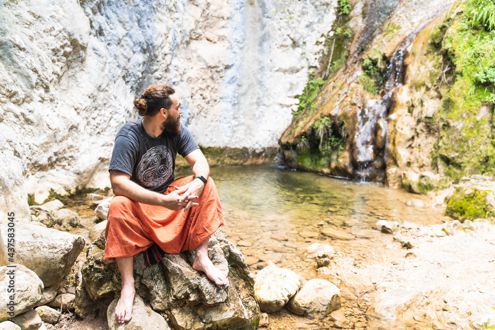 Caucasian man with beard and long hair looking at the sky while sitting on a rock next to a waterfall and a lake in the Sierra de Tramuntana, Palma de Mallorca, Spain (Copyspace) Caucasian man with beard and long hair looking at the sky while sitting on a rock next to a waterfall and a lake in the Sierra de Tramuntana, Palma de Mallorca, Spain (Copyspace)