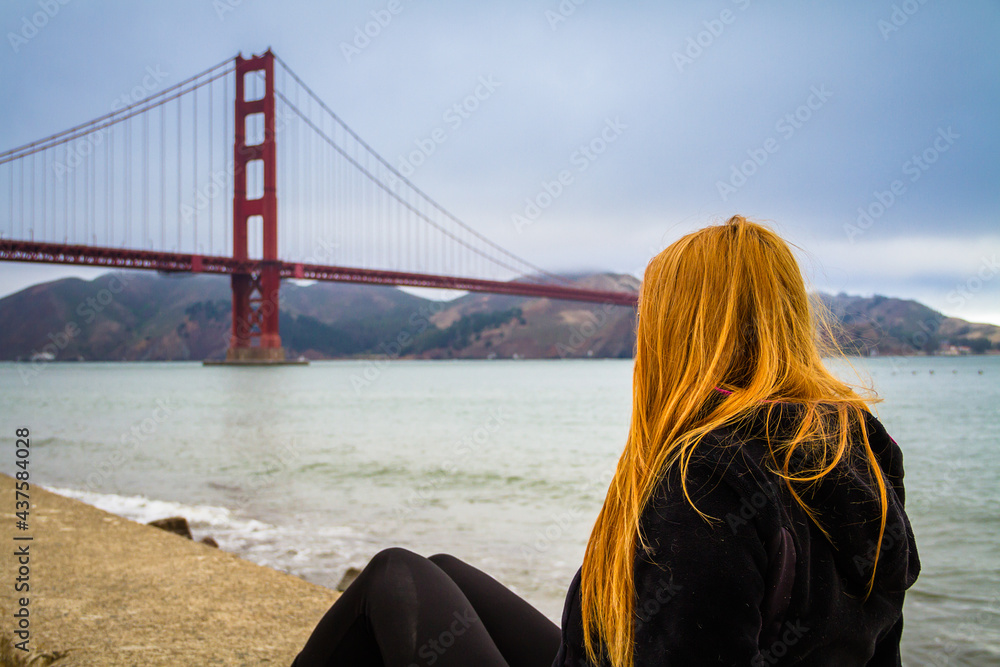A young woman with red long hair sitting by San Francisco Bay watching the Golden Gate Bridge