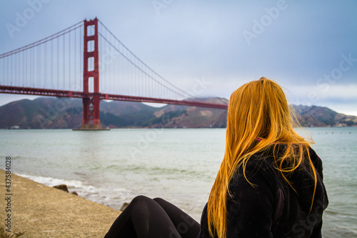 A young woman with red long hair sitting by San Francisco Bay watching the Golden Gate Bridge