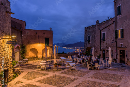 Borgio Verezzi, Italy. May 22th, 2021. Piazza Sant'Agostino at night with people sitting outdoors at a restaurant table. In the distance the view on the Ligurian Riviera in the background.