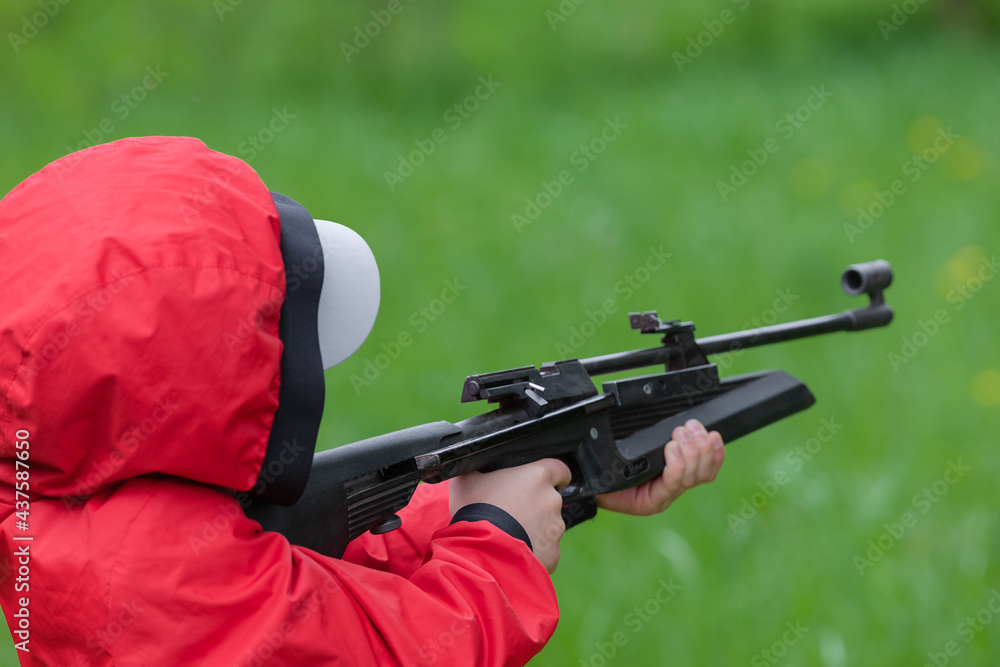 A boy in a red windbreaker and a cap is holding an air gun. The concept of weapons and children, training in shooting. A boy in a red windbreaker and a cap is holding an air gun. The concept of weapons and children, training in shooting.