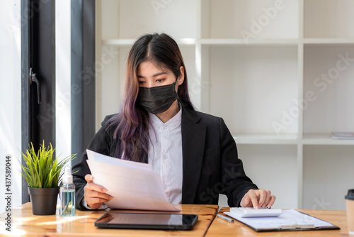 Young Asian businesswoman working using a calculator and document with a laptop at the office wear a black mask to protect against germs.