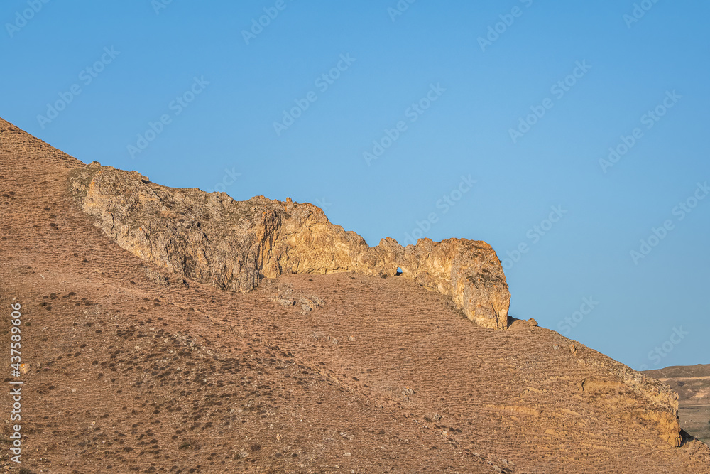 A rock of bizarre shape in a desert landscape against a blue sky.