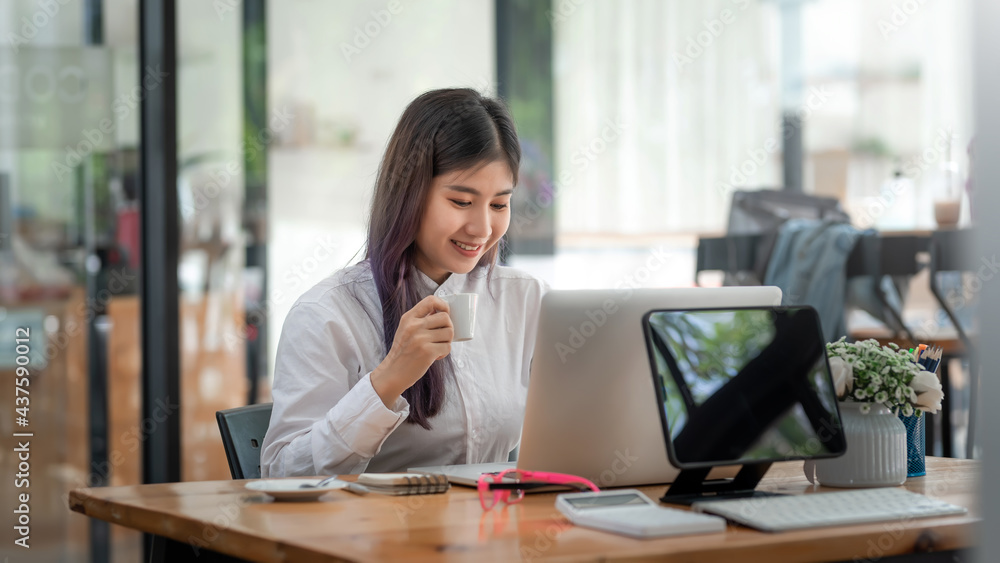 Smiling young Asian woman working on laptop and drinking coffee at office. Smiling young Asian woman working on laptop and drinking coffee at office.