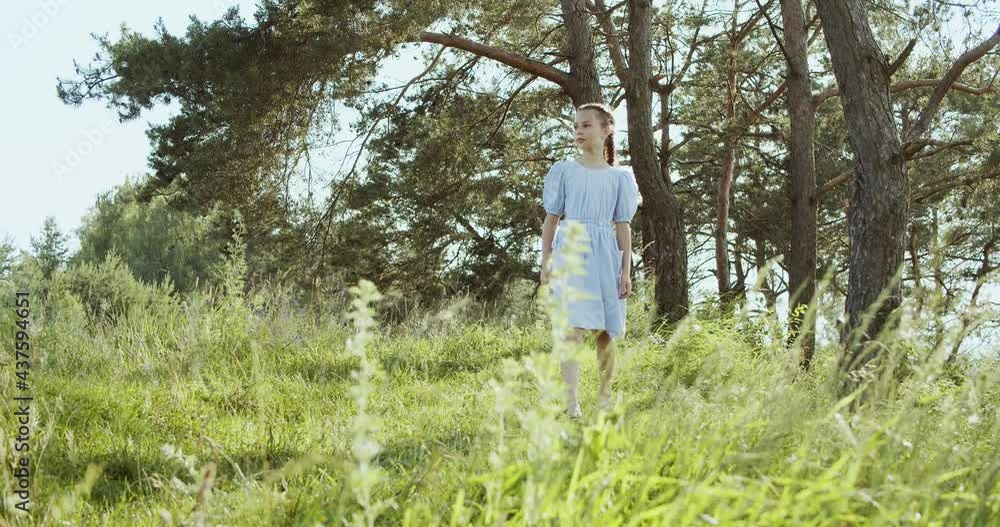 A cheerful teenage girl with braided hair walks in a clearing between green trees in summer forest. A cheerful teenage girl with braided hair walks in a clearing between green trees in summer forest.