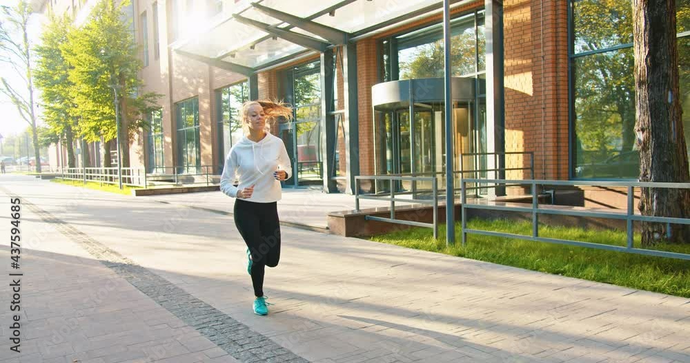 Caucasian young beautiful happy sportswoman running in sunny town in good mood, morning exercises. Joyful pretty female jogging outdoors listening to music in earphones. Sport lifestyle concept Caucasian young beautiful happy sportswoman running in sunny town in good mood, morning exercises. Joyful pretty female jogging outdoors listening to music in earphones. Sport lifestyle concept