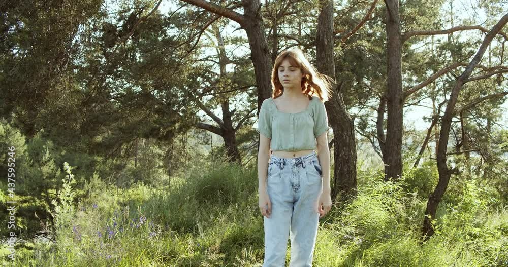 A cheerful teenage girl with curly hair stands in a clearing between green trees in summer forest. A cheerful teenage girl with curly hair stands in a clearing between green trees in summer forest.