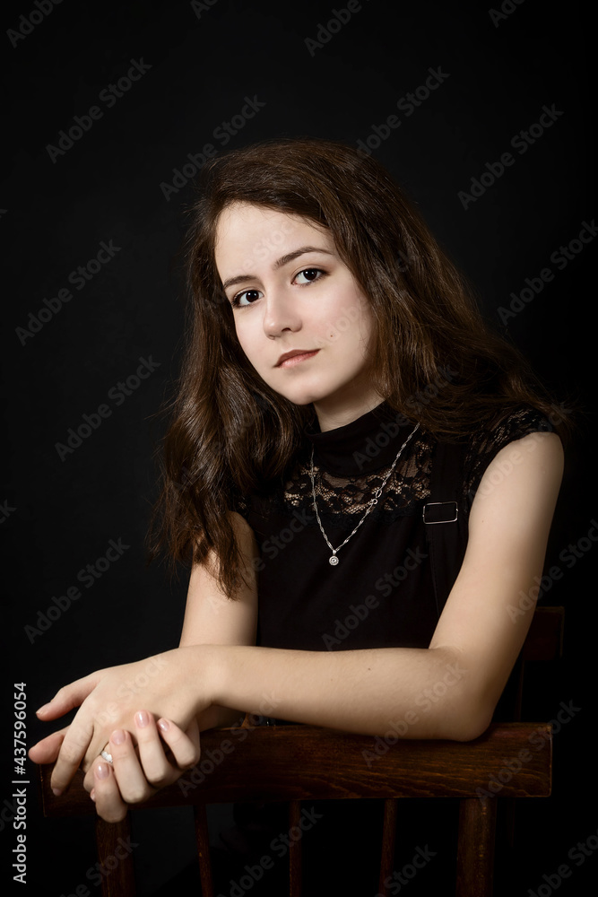 Portrait of a smiling beautiful young brown-haired woman with wavy hair, in black clothes on a black background, sitting on a chair; vertical Portrait of a smiling beautiful young brown-haired woman with wavy hair, in black clothes on a black background, sitting on a chair; vertical