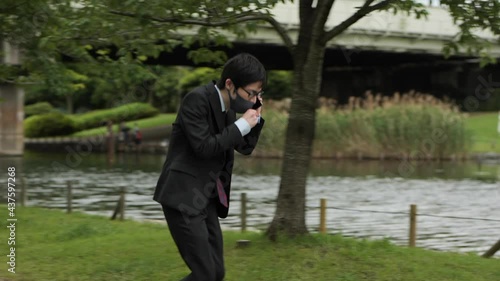 コロナ禍に川原でビジネスパーソンがマスクをして電話をしながら咳込む