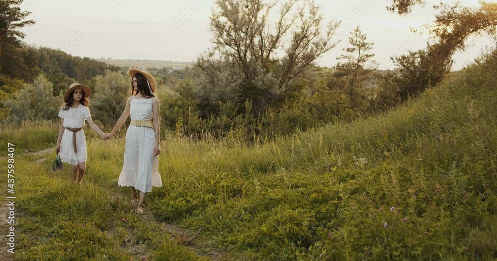 Two girl teenagers in white dresses and straw hats with flowers bouquet walking on nature at sunny summer day Two girl teenagers in white dresses and straw hats with flowers bouquet walking on nature at sunny summer day