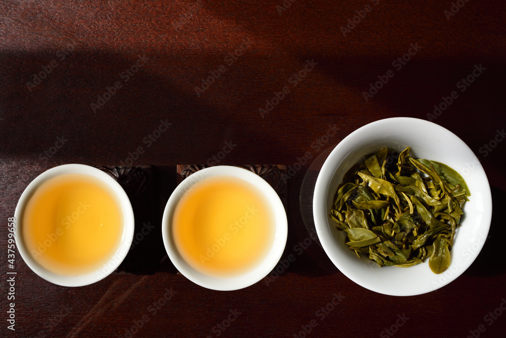 Still life and on top shot of two Chinese tea cups with golden tea and a teapot with fresh green tea leaves, side by side on a brown table
