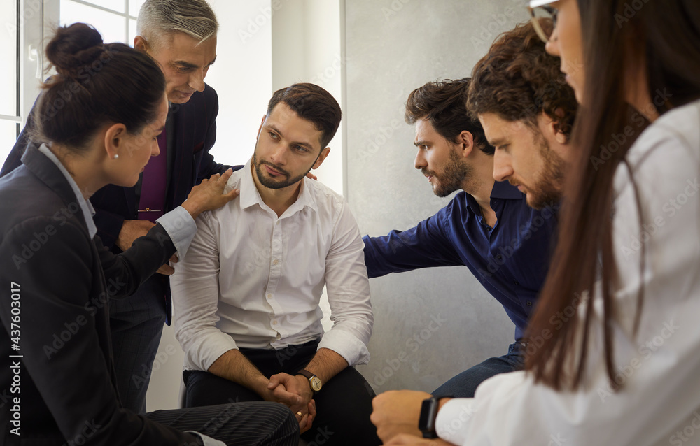Group of different people sitting in a circle supporting and comforting a sad upset young man. Concept of psychological help, group therapy and overcoming problems together Group of different people sitting in a circle supporting and comforting a sad upset young man. Concept of psychological help, group therapy and overcoming problems together