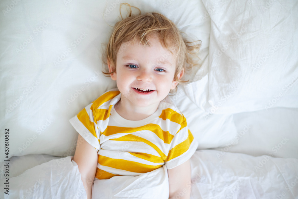 Top view of cheerful little boy in white bed early in the morning. Portrait of a handsome boy awakened