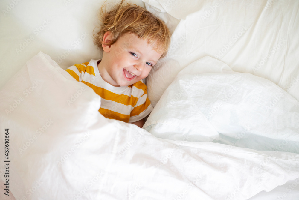 Top view of cheerful little boy in white bed early in the morning. Portrait of a handsome boy awakened Top view of cheerful little boy in white bed early in the morning. Portrait of a handsome boy awakened