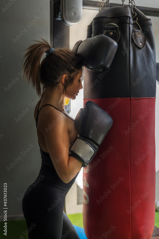 a full-length girl rests during a boxing training session. she leans on the training bag. the light cuts out her silhouette.