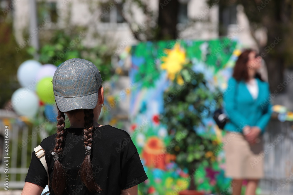Teenager girl with pigtails in a baseball cap in a black T-shirt on the street against a background of colorful balloons and festive decorations.Portrait of a child close-up view from the back Teenager girl with pigtails in a baseball cap in a black T-shirt on the street against a background of colorful balloons and festive decorations.Portrait of a child close-up view from the back