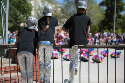 Three middle school girls in the same uniform black T shirt and silver baseball cap and leggings with long hair with a pigtail watch the performance at a children's party sitting on a fence outdoors