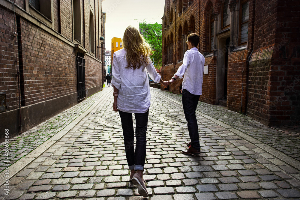 Zwei junge Leute, eine Frau und ein Mann gehen durch die Stadt Hand an der Hand spazieren. Der Weg ist gepflastert, die Straße ist eng. Sie haben weiße Hemd an. Sie hat lange, blonde Haare