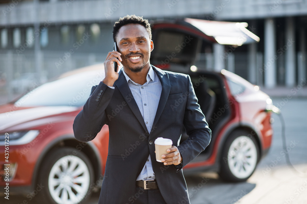Busy african man in formal suit having mobile conversation while his luxury red car is charging. Businessman solving working issues on smartphone while waiting for electric auto. Busy african man in formal suit having mobile conversation while his luxury red car is charging. Businessman solving working issues on smartphone while waiting for electric auto.