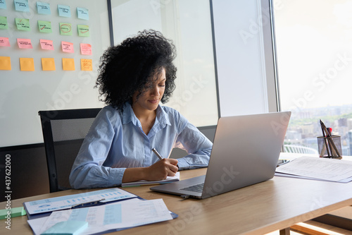 Tableau sur toile Young African American focused female ceo data analyst businesswoman sitting at desk with laptop writing notes near panoramic window in contemporary corporation office