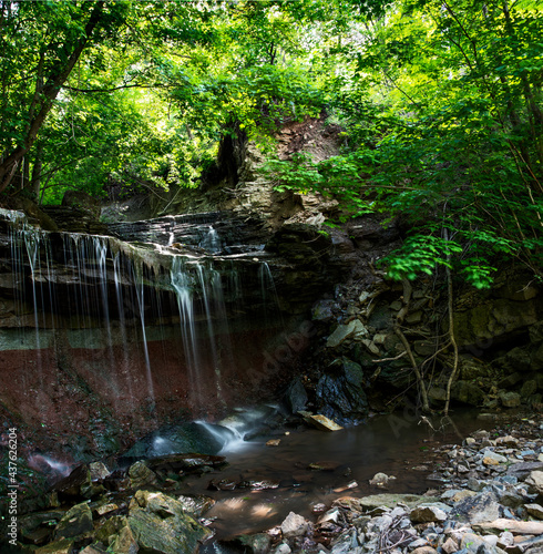 A small waterfall cascades down a rocky hill through a lush green forest into a pool of rocks below.