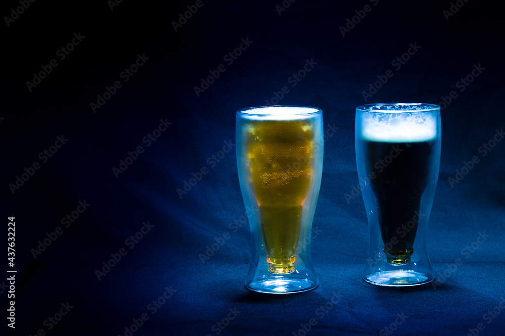 Two glasses of cold and refreshing light and dark beer on a blue cloth and a blue background with a dim light on them Two glasses of cold and refreshing light and dark beer on a blue cloth and a blue background with a dim light on them