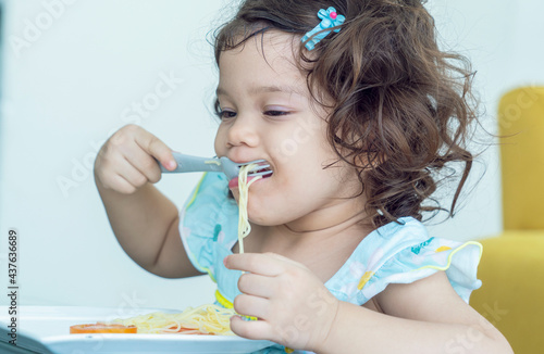 Konstfotografi Hungry little daughter sits and eats spaghetti noodles with a spoon and eats deliciously in the kitchen at home
