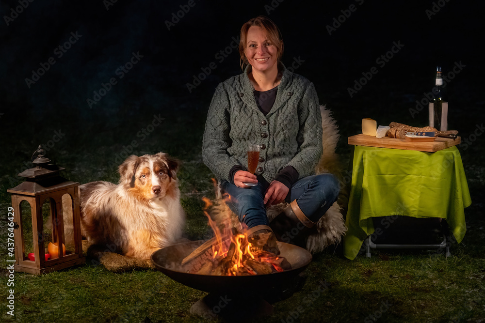 Young woman drinks red wine by the campfire in the forest. Her Australian shepperd sits next to her on a rug in the snowy grass. Enjoying food and drink in leisure time in winter