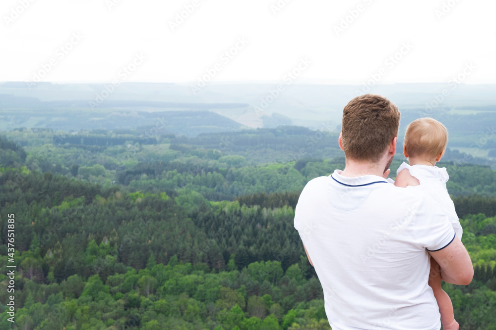 Handsome caucasian man with baby in hands look at beautiful panoramic view on green forest in Kislovodsk park. Father and kid travel together. Spending time with child on vacation concept. Back view