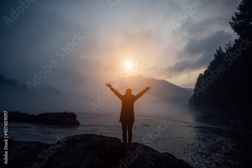 Blue evening mist over mountain river on the background of wooded hills and overcast sky. Man stands in the fog, hands up, looking at the setting sun. Woman's appeal to the power of nature.
