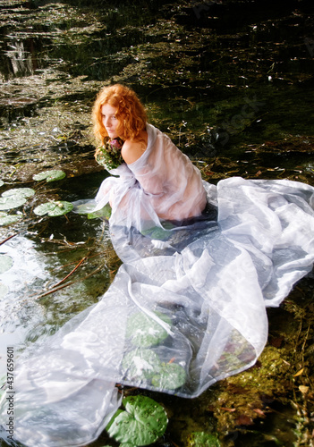 young sexy woman, redhead Ophelia, with curly red hair sits joyfully, smiling, with white dress and a withered bouquet of flowers in the water in the lake, copy space photo