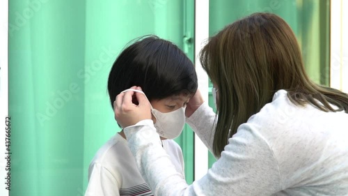 Close-up of an Asian mother putting a protective medical face mask on her little son outdoors.