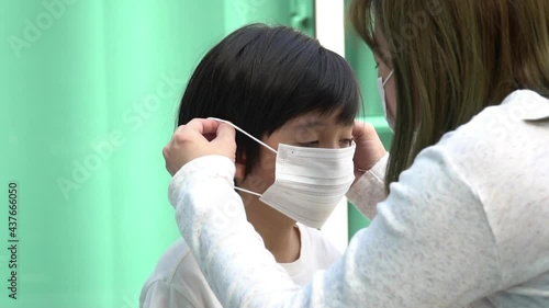 Close-up of an Asian mother putting a protective medical face mask on her little son outdoors.