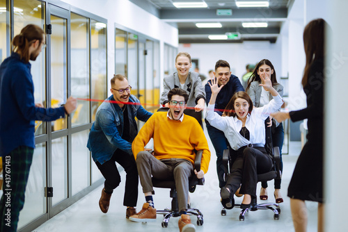 Young colleagues group having fun together, riding on chairs in office, diverse excited office workers enjoying break, laughing, engaged funny activity, celebrating corporate success.