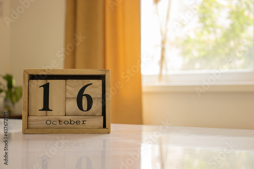 Wooden blocks of the calendar represents the date 16 and the month of October on the background of a window, curtain and a plant.