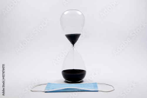 An hourglass against a white background with a soft focus medical mask laying in front of it.