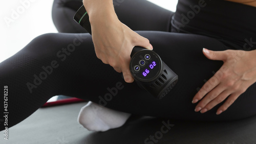 Young fit woman practicing self-massage technique for buttocks applying therapeutic percussive massage gun sitting on pilates mat at home or studio interior.