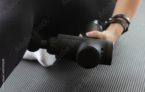 Young fit woman practicing self-massage technique for buttocks applying therapeutic percussive massage gun sitting on pilates mat at home or studio interior.