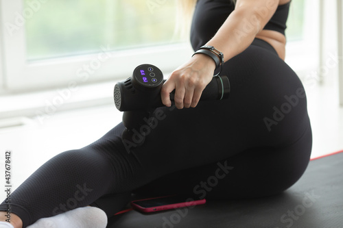 Young fit woman practicing self-massage technique for buttocks applying therapeutic percussive massage gun sitting on pilates mat at home or studio interior.