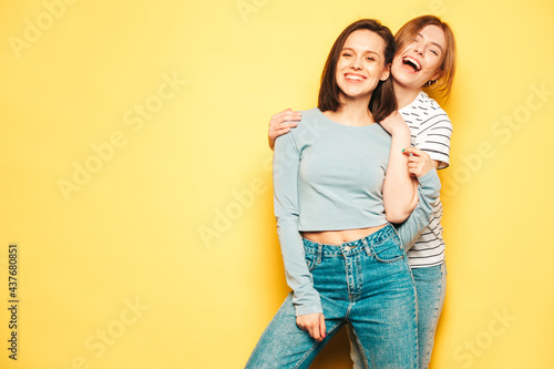 Two young beautiful smiling hipster female in trendy summer white t-shirt and jeans clothes.Sexy carefree women posing near yellow wall in studio.Positive and cheerful models