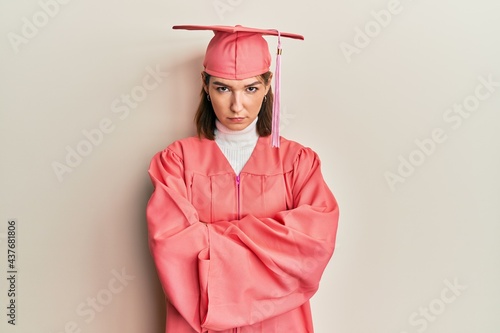 Young caucasian woman wearing graduation cap and ceremony robe skeptic and nervous, disapproving expression on face with crossed arms. negative person.