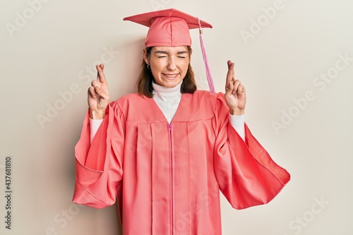 Young caucasian woman wearing graduation cap and ceremony robe gesturing finger crossed smiling with hope and eyes closed. luck and superstitious concept.