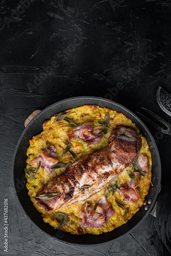 Grilled pork fillet and mash potatoe gratin with sage and prosciutto , on frying cast iron pan , with barbeque knife and meat fork, on black stone background, top view flat lay