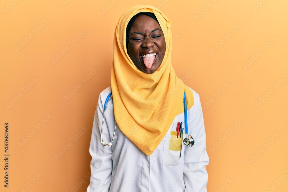 Beautiful african young woman wearing doctor uniform and hijab sticking tongue out happy with funny expression. emotion concept. Beautiful african young woman wearing doctor uniform and hijab sticking tongue out happy with funny expression. emotion concept.