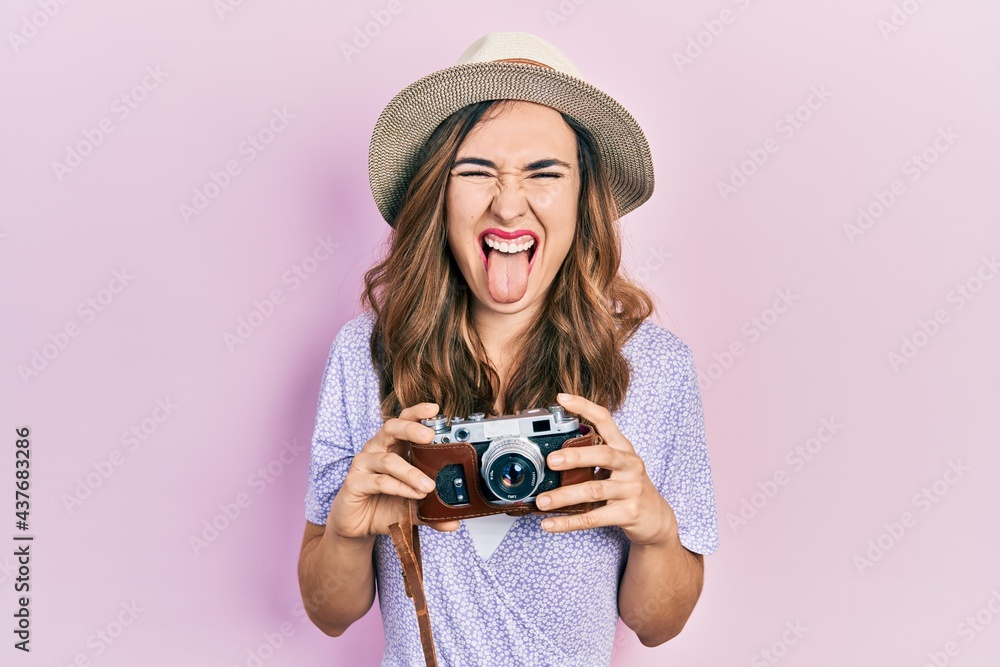 Young hispanic girl wearing summer hat holding vintage camera sticking tongue out happy with funny expression.